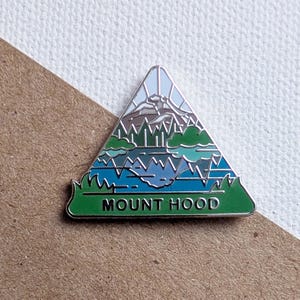 May include: A triangular enamel pin featuring a mountain landscape with the text "MOUNT HOOD". The design includes a mountain peak, trees, and a lake, with shades of blue, green, and brown. The pin has a silver border and a green base.