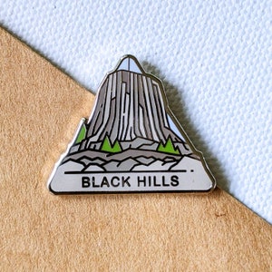 May include: A silver enamel pin featuring a stylised illustration of a mountain with a white peak and green trees. The text "BLACK HILLS" is printed below the mountain.