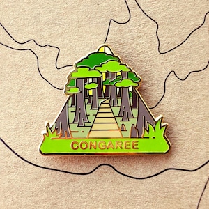 May include: A gold-colored enamel pin with a green and brown illustration of a cypress swamp with a path leading through it. The text "CONGAREE" is printed in gold on the bottom of the pin.