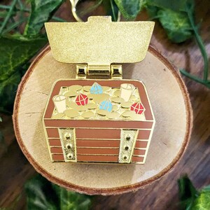 K&ouml;nnte beinhalten: Goldfarbene Schatzkiste-Anstecknadel mit braunem Holzschrank-Design. Die Truhe ist mit Goldm&uuml;nzen, roten und blauen Edelsteinen und kleinen wei&szlig;en Bechern gef&uuml;llt. Die Nadel hat einen goldfarbenen Verschluss oben.