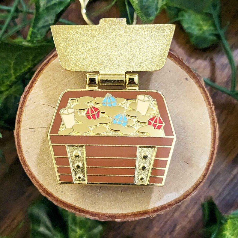 K&ouml;nnte beinhalten: Goldfarbene Schatzkiste-Anstecknadel mit braunem Holzschrank-Design. Die Truhe ist mit Goldm&uuml;nzen, roten und blauen Edelsteinen und kleinen wei&szlig;en Bechern gef&uuml;llt. Die Nadel hat einen goldfarbenen Verschluss oben.