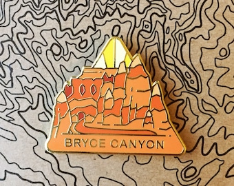 May include: A gold-colored enamel pin featuring a stylized depiction of Bryce Canyon National Park in Utah. The pin is orange, red, and yellow with a black outline. The text "BRYCE CANYON" is printed in black on the bottom of the pin.