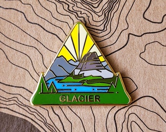 May include: A gold-colored enamel pin shaped like a triangle with a mountain scene inside. The pin features a mountain range with a lake in the foreground and a sun rising behind the mountains. The text "GLACIER" is printed in black at the bottom of the pin.