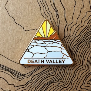 May include: Gold-coloured enamel pin shaped like a triangle with a white background. The pin features a stylised image of a sunset over a cracked desert landscape. The text "DEATH VALLEY" is printed in black at the bottom of the pin.