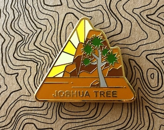 May include: A gold-colored enamel pin with a triangular shape. The pin features a stylized Joshua Tree with a yellow sun rising behind it. The text "Joshua Tree" is printed in black at the bottom of the pin.
