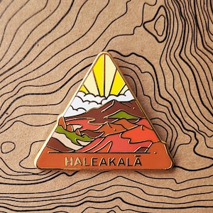 May include: A gold triangle-shaped enamel pin with a colorful illustration of a mountain range and a sun rising over the mountains. The text "HALEAKALĀ" is printed in black at the bottom of the pin.