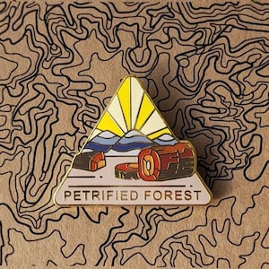 May include: A triangular enamel pin with a gold outline and a pink background. The pin features a stylized sunrise with a blue mountain range and two brown logs. The text "Petrified Forest" is printed in gold letters at the bottom.