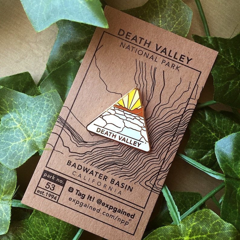 May include: Gold enamel pin depicting a stylised mountain range with a sunrise over a desert landscape. The pin reads "Death Valley" and is on a brown card with the text "Death Valley National Park" and "Badwater Basin, California".