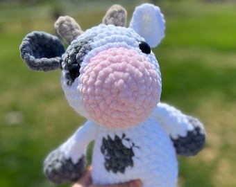 May include: A plush cow toy made of white, gray, and light pink yarn. The stuffed animal features a white body with gray patches, a light pink snout, two small gray horns, and black plastic eyes. The texture is soft and crocheted throughout.