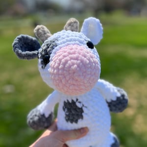 May include: A crocheted plush toy cow with white and gray fur, pink nose, and black eyes. The cow is being held by a hand wearing a gray bracelet.
