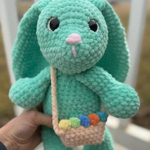 Puede incluir: Un conejito de crochet verde con la nariz rosa y ojos negros brillantes. El conejito lleva una pequeña bolsa de crochet con pompones arcoíris.