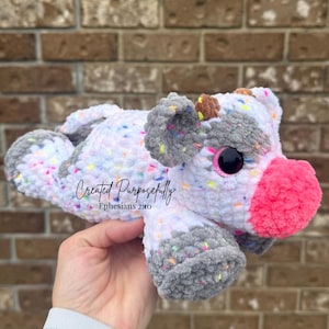 Può includere: Un giocattolo di peluche di mucca all'uncinetto bianco e grigio con accenti rosa e spolverata colorata. Il giocattolo ha grandi occhi neri e un naso marrone. Il giocattolo è tenuto in una mano e il testo "Created Purposefully Ephesians 2:10" è visibile.