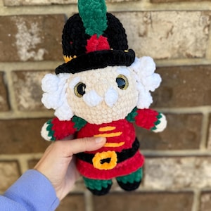 May include: A handmade crochet nutcracker doll. The doll has a black hat with a green feather and red accents. It wears a red jacket with gold details and a black belt. The doll has a cream-colored face with white hair and gold eyes.