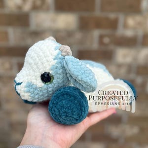 Può includere: Un agnello all'uncinetto fatto a mano in bianco e azzurro. L'agnello ha un corpo blu teal e un occhio verde. Il testo "CREATED PURPOSEFULLY EPHESIANS 2:10" è sul lato destro dell'agnello.