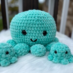 May include: A turquoise crocheted octopus plushie with three smaller octopus companions. The large octopus has black button eyes and a smiling mouth. The plushies are made of soft, textured yarn and sit on a white, fluffy surface.