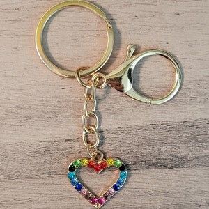 May include: Gold-toned keychain with a heart-shaped charm. The charm is outlined with colorful rhinestones in a rainbow pattern. The keychain includes a split ring, a lobster clasp, and a short chain.
