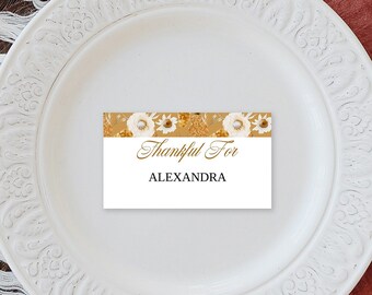 Puede incluir: Un plato blanco con borde decorativo contiene una tarjeta de mesa que dice "Thankful For" y "Alexandra" en letras doradas. La tarjeta de mesa tiene un diseño floral. La mesa incluye una cuchara dorada, velas y flores secas.