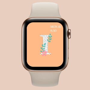 May include: A white Apple Watch with a gold band and a peach-colored watch face. The watch face displays the date "MON 10" and the time "8:30". The watch face also features a large, stylized letter "I" with black polka dots and a floral design.