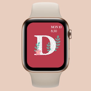 May include: A gold Apple Watch with a white band displays a red watch face with a white letter "D" with black polka dots and green leaves. The watch face also displays the date "MON 10" and the time "8:30".