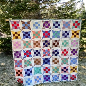 May include: A colorful handmade quilt with a repeating pattern of squares and circles. The quilt features a variety of fabrics in shades of red, blue, yellow, green, and purple. The quilt is hanging outside, with a tree in the background.