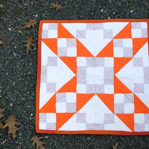 May include: A white and orange patchwork quilt with a star pattern. The quilt is made of squares and triangles and has a white background with orange stars.