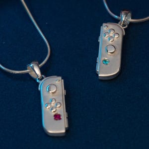 May include: Two silver pendant necklaces featuring a video game controller design. One pendant has a red gem, the other a blue gem. The pendants hang from delicate silver chains, set against a dark blue background.