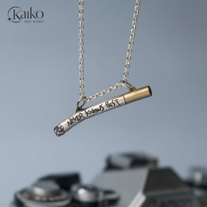 Puede incluir: Un collar de plata con un colgante con forma de cigarrillo que dice "NEVER KNOWS BEST" en letras negras. El colgante tiene una punta dorada.