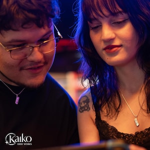 May include: Two people are looking at something, each wearing a silver necklace with a small, rectangular pendant. The pendants appear to be a video game controller. The image has a blue and purple color scheme. The Kaiko Geek Works logo is in the bottom left corner.