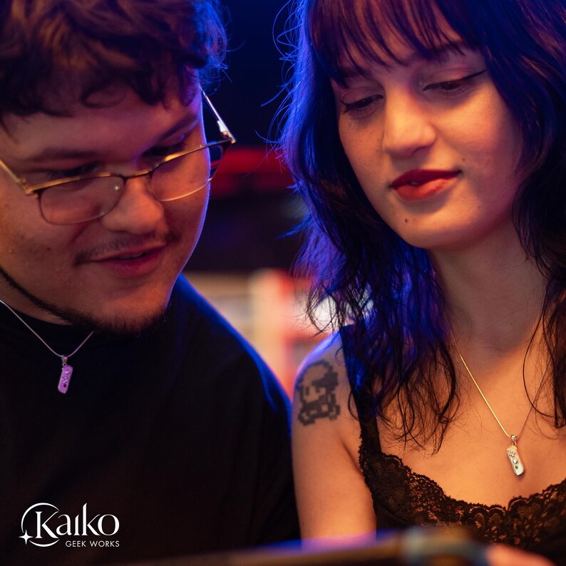 May include: Two people are looking at something, each wearing a silver necklace with a small, rectangular pendant. The pendants appear to be a video game controller. The image has a blue and purple color scheme. The Kaiko Geek Works logo is in the bottom left corner.