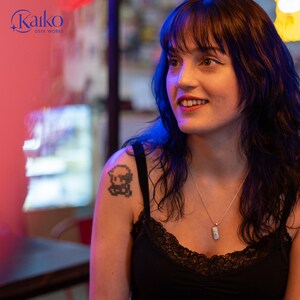 May include: A woman wearing a black tank top with lace trim and a silver necklace with a small pendant. A pixelated tattoo is visible on her shoulder. The Kaiko Geek Works logo is in the upper left corner.