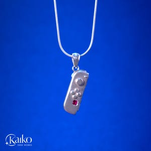 May include: A silver pendant necklace featuring a miniature game controller design. The controller has button details and a red gemstone accent. The necklace hangs on a thin silver chain against a blue background.