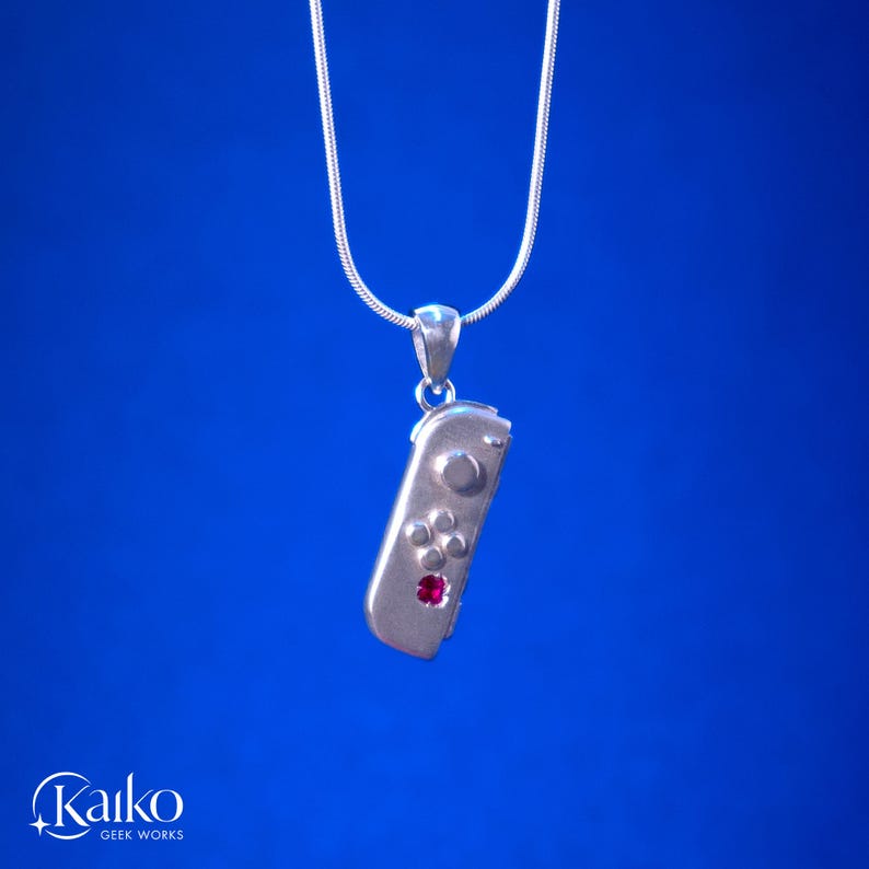 May include: A silver pendant necklace featuring a miniature game controller design. The controller has button details and a red gemstone accent. The necklace hangs on a thin silver chain against a blue background.