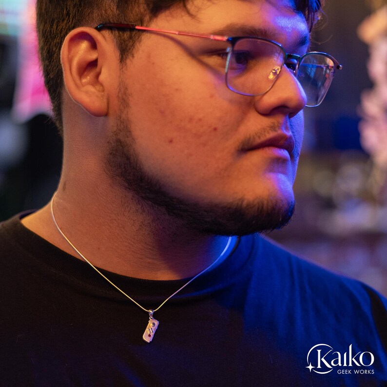 May include: A silver pendant necklace with a rectangular charm. The charm has a detailed design with small, colorful accents. The necklace is worn on a black shirt. The Kaiko Geek Works logo is visible in the bottom right corner.