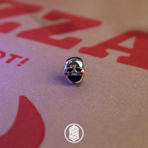 May include: A small, silver-colored charm in the shape of a stylized mask. The mask has black and blue eye details, a wide black mouth, and red accents. The charm is set against a background with red text and a logo.