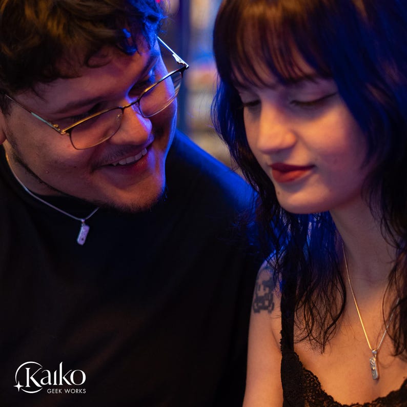 May include: A photograph of two people, both wearing silver necklaces with rectangular pendants. The man is wearing glasses and a black shirt. The woman is wearing a black top. The logo "Kaiko Geek Works" is in the bottom left corner.