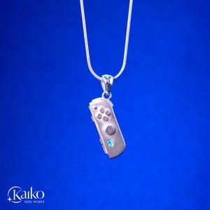 May include: A silver pendant necklace featuring a miniature game controller design. The controller has button details and a small blue gemstone. The necklace hangs on a silver chain against a blue background.