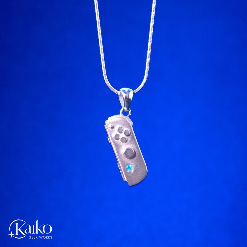 May include: A silver pendant necklace featuring a miniature game controller design. The controller has button details and a small blue gemstone. The necklace hangs on a silver chain against a blue background.