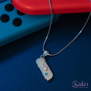 May include: Silver-colored pendant necklace featuring a miniature game controller design. The controller has detailed button and joystick representations, with a small blue gem accent. The necklace is displayed on a dark blue surface.