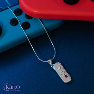May include: A silver pendant necklace featuring a miniature game controller design with a small red gemstone. The pendant hangs on a delicate silver chain. The background includes a blue and red game controller.