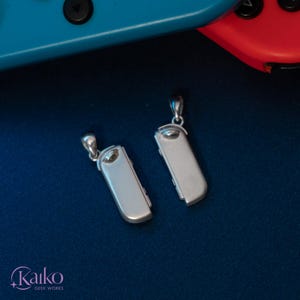 May include: Two silver-colored pendant necklaces, each shaped like a game controller. The pendants are rectangular with rounded edges and a small loop at the top for the chain. The background is a dark blue surface, with a blue and red game controller in the background.