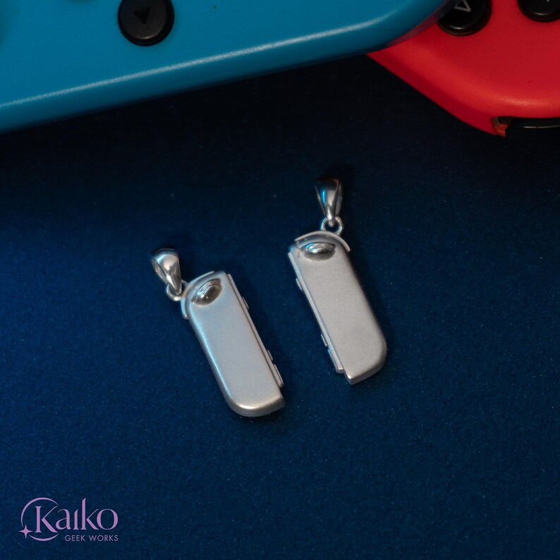 May include: Two silver-colored pendant necklaces, each shaped like a game controller. The pendants are rectangular with rounded edges and a small loop at the top for the chain. The background is a dark blue surface, with a blue and red game controller in the background.