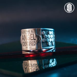 Könnte beinhalten: Ein silberner Ring mit den eingravierten Texten "BLOOD TYPE: BLUE" und "ANGEL IDENTIFIED". Der Ring hat ein Wabenmuster auf einer Seite und den Text "MAGI - 1-2-3" darunter. Der Ring liegt auf einer reflektierenden Oberfläche.