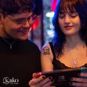May include: Two people are looking at a Nintendo Switch game console. Both are wearing silver necklaces with rectangular pendants. One person has a pixelated tattoo on their shoulder. The Kaiko Geek Works logo is in the bottom left corner.