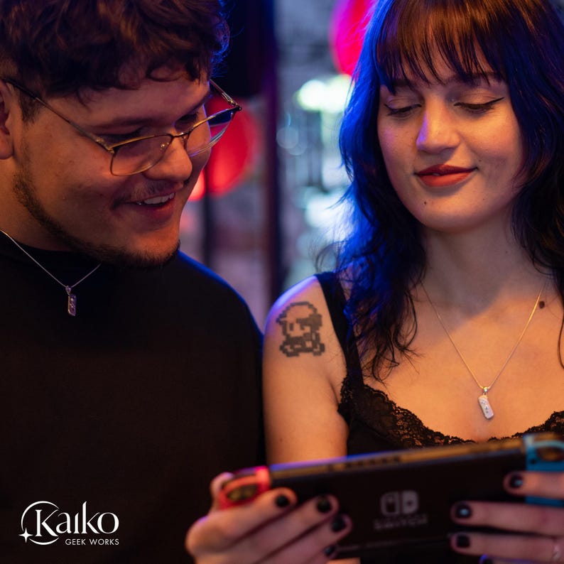 May include: Two people are looking at a Nintendo Switch game console. Both are wearing silver necklaces with rectangular pendants. One person has a pixelated tattoo on their shoulder. The Kaiko Geek Works logo is in the bottom left corner.