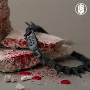 May include: A black, articulated necklace with a stylized cat face pendant and spiky segments. The necklace is displayed on a rough, reddish-brown stone surface with red droplets and small white stones.