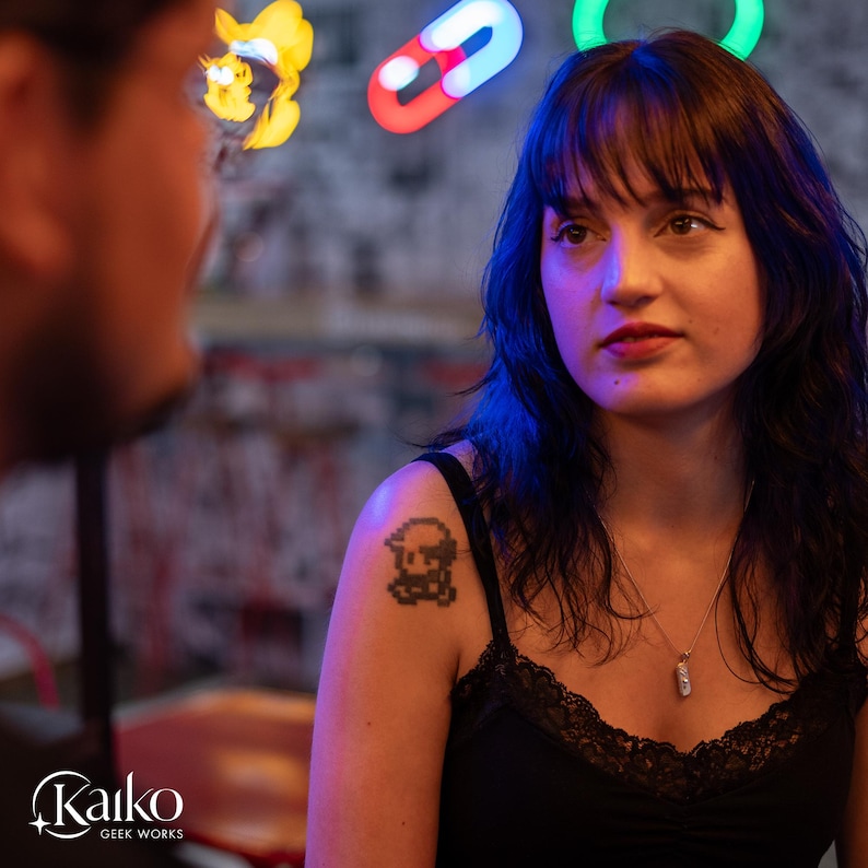 May include: A woman with dark hair and a black tank top with a lace trim. She has a pixelated tattoo on her shoulder. The image has a neon light design in the background. The Kaiko Geek Works logo is in the bottom left corner.