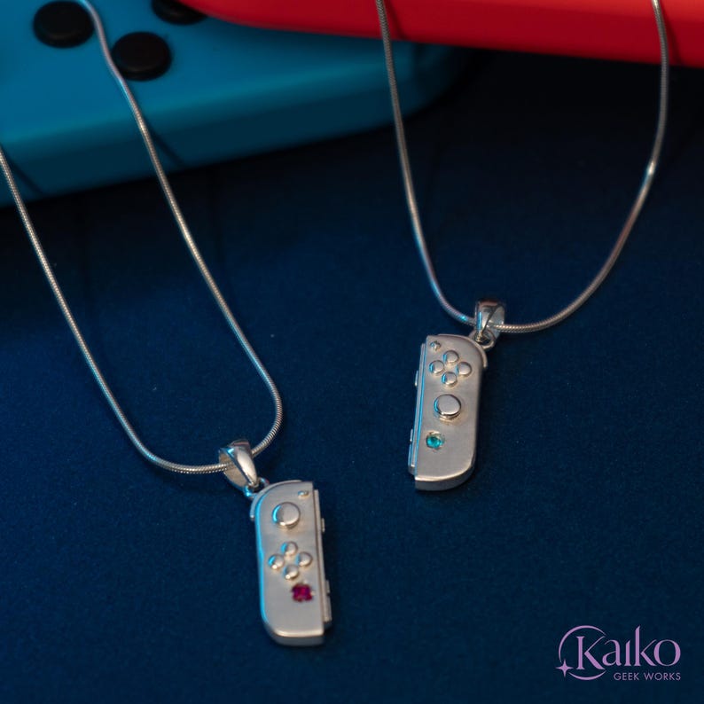May include: Two silver pendant necklaces featuring a video game controller design. One pendant has a red gem, the other a blue gem. The pendants hang from delicate silver chains, set against a dark blue background.