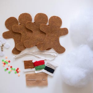 May include: A craft kit with brown felt gingerbread man shapes, white stuffing, spools of thread, beads, and a needle. The kit includes various craft supplies for creating handmade decorations or ornaments.
