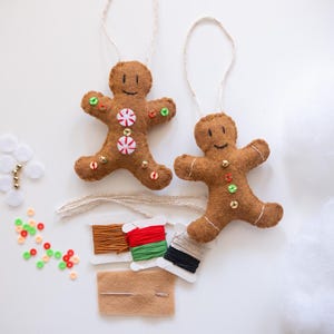 May include: Two brown felt gingerbread man ornaments with white string hangers. One has candy embellishments. Craft supplies include thread, beads, and a needle. Festive holiday decor.