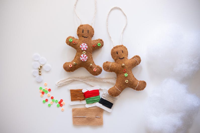 May include: Two brown felt gingerbread man ornaments with white string hangers. One has candy embellishments. Craft supplies include thread, beads, and a needle. Festive holiday decor.